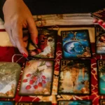Mãos segurando cartas de Baralho Cigano com um notebook ao fundo exibindo um curso online